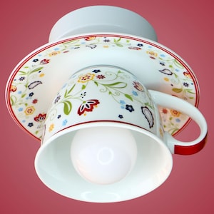Könnte beinhalten: Eine dekorative Deckenleuchte in Form einer Tasse und Untertasse. Die weiße Keramik-Tasse und Untertasse sind mit einem Blumenmuster in Rot, Gelb, Blau und Grün verziert. Eine Glühbirne befindet sich in der Tasse, und die Leuchte hat eine weiße Basis.