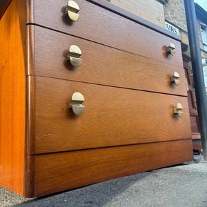 May include: A wooden chest of drawers with a warm brown finish, featuring three horizontal drawers. Each drawer is equipped with two semi-circular, metallic gold-toned handles positioned on the left and right sides of the drawer fronts.