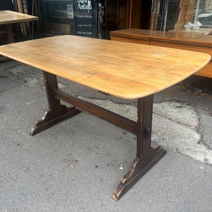 May include: A rectangular wooden table featuring a light brown, natural wood grain top with rounded corners. The table is supported by a dark brown trestle base with two vertical uprights connected by a horizontal stretcher bar near the floor.