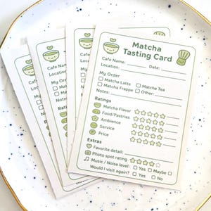 May include: A stack of Matcha Tasting Cards on a white plate. Each card features a green design with a bowl of matcha and text fields for cafe name, location, order, and ratings for flavor, ambiance, and service. The cards are designed for cafe reviews.