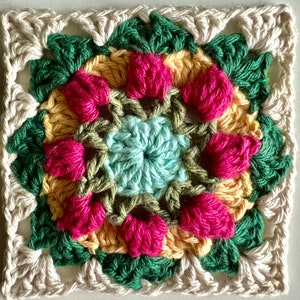 May include: A crocheted square with a floral design in shades of pink, yellow, green, and blue. The center of the square features a smaller circle with a light blue center.