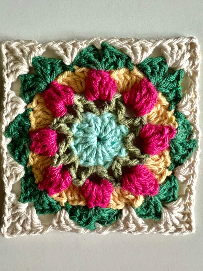 May include: A crocheted square with a floral design in shades of pink, yellow, green, and blue. The center of the square features a smaller circle with a light blue center.