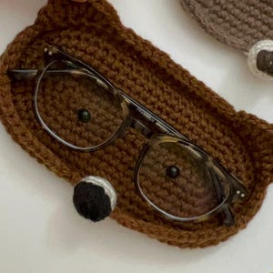 May include: Brown crocheted glasses case shaped like a bear with a black nose and glasses inside.
