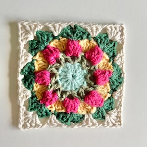 May include: A crocheted square with a colorful floral design in shades of pink, yellow, green, and blue. The center of the square features a smaller, light blue circle.