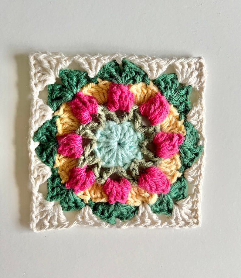 May include: A crocheted square with a colorful floral design in shades of pink, yellow, green, and blue. The center of the square features a smaller, light blue circle.