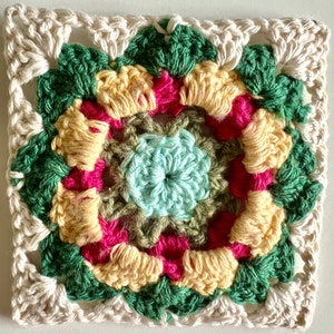 May include: A crocheted square with a multi-colored floral design in shades of green, yellow, pink, and light blue. The square is surrounded by a white crocheted border.