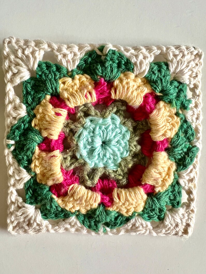 May include: A crocheted square with a multi-colored floral design in shades of green, yellow, pink, and light blue. The square is surrounded by a white crocheted border.