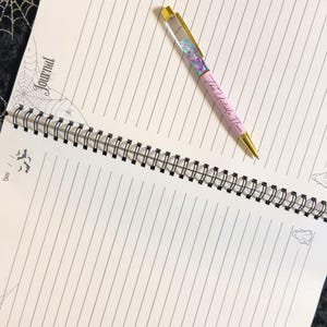 May include: A pink pen with gold accents and the text "The Unstable Flower" rests on an open spiral-bound notebook. The pen's barrel is filled with glitter and small, colorful shapes. The notebook has lined pages and decorative Halloween-themed illustrations.