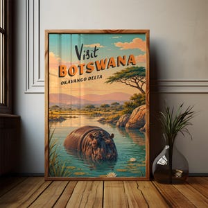 Op de afbeelding: Een ingelijste reisposter met een nijlpaard in de Okavangodelta met de tekst Visit BOTSWANA OKAVANGO DELTA. De illustratie toont een zonsondergang, acacia's, rotsen en waterlelies in een rivier, geplaatst in een houten lijst.