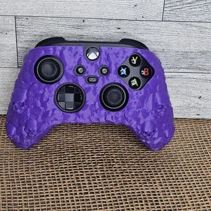 May include: Purple gaming controller with a textured skull design. The controller features black buttons and directional pad. The Xbox logo is visible in the center. A unique accessory for gamers.
