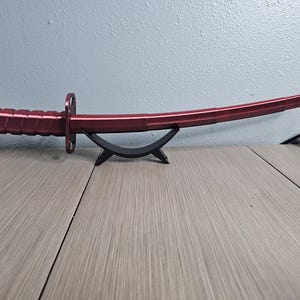 May include: A red katana sword with a curved blade, resting on a black stand. The handle has a textured grip, and the blade is a solid, vibrant red. The sword is displayed on a wooden surface.