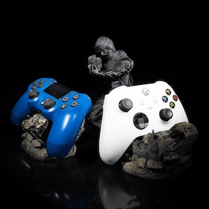 May include: A blue PlayStation controller and a white Xbox controller rest on textured, gray bases. A small, dark figurine of a person holding a weapon stands between them against a black background. The controllers have black buttons.