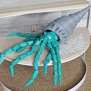 May include: A teal and gray articulated sea creature toy with a large, spiraled shell. The toy has multiple jointed appendages and a cartoonish face. It is displayed on a small, round wooden shelf with a fern design.