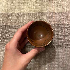 May include: A small, brown ceramic cup with a dark brown glaze. The cup is empty and has a slightly rounded shape.
