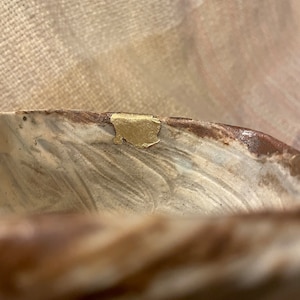 Puede incluir: Primer plano de un cuenco de cerámica con un esmalte marmoleado marrón y blanco. El cuenco tiene un acento dorado en el borde.