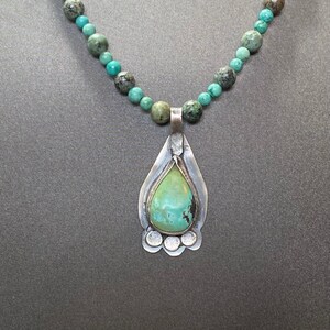 May include: A turquoise beaded necklace with a silver pendant. The pendant has a teardrop-shaped turquoise stone set in a silver frame. The necklace features turquoise and gray-green beads.