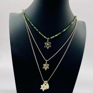 May include: Three silver necklaces with Star of David pendants. The top necklace is made of green and blue beads. The middle necklace has a pendant with a small eye in the centre. The bottom necklace has a pendant with a larger eye in the centre.