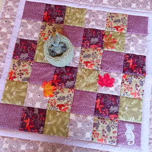 May include: A patchwork quilt with squares of purple, olive green, and patterned fabrics. The quilt features appliqued autumn leaves and a small stuffed animal in a green nest. The quilt is bordered with a white edge.