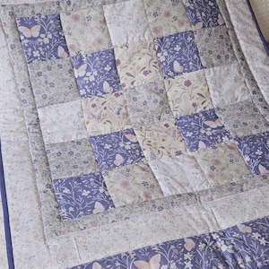 May include: A patchwork quilt in lavender, white, and grey, featuring a floral and butterfly design. The quilt has a lavender border. The item is handmade, with a cosy, floral theme.