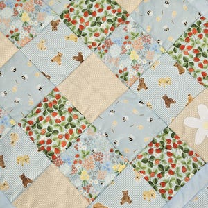 May include: A patchwork quilt with a light blue border. The quilt features squares with teddy bears, strawberries, bees, and floral patterns in shades of blue, beige, and red. A white daisy is appliquéd on the quilt.