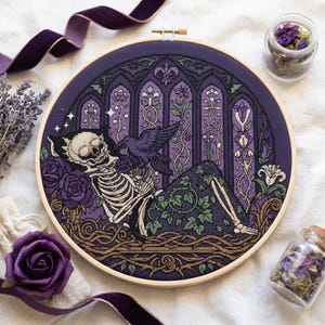 May include: A circular embroidery hoop containing a cross-stitch design of a human skeleton reclining amidst purple roses and green ivy. A black raven sits on the skeleton's chest before a backdrop of arched gothic windows in shades of purple.