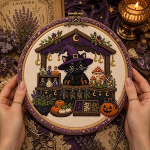 Peut inclure: Un tambour à broder circulaire représentant un chat noir portant un chapeau de sorcière violet derrière un étal en bois. La scène inclut des fioles colorées, des champignons, des citrouilles, des herbes suspendues et une citrouille d'Halloween.