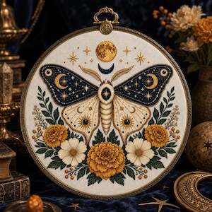 Könnte beinhalten: Ein runder Stickrahmen mit einem Kreuzstichmotiv eines Nachtfalters, dessen dunkelblaue Flügel mit goldenen Mondsicheln und Sternen verziert sind, umgeben von gelben und weißen Blumen auf einem weißen Stoffhintergrund.