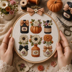 May include: A circular wooden embroidery hoop containing a cross-stitch pattern on white fabric. The design features six distinct motifs: a black jar with a plaid bow, a pumpkin with white flowers, a striped jar, and three vases with floral arrangements.
