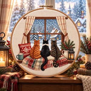 May include: A circular cross-stitch embroidery hoop featuring three cats sitting on a windowsill looking out at a snowy winter landscape. The scene includes a red patterned pillow, a plaid blanket, a mug, and a small lantern on the windowsill.