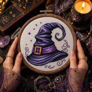 Peut inclure: Un tambour à broder circulaire en bois contenant un motif au point de croix représentant un chapeau de sorcière violet avec une boucle dorée. Le chapeau est centré sur un tissu blanc, entouré de petites étoiles violettes et de volutes.