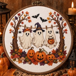 May include: A circular cross-stitch embroidery in a wooden hoop featuring three white ghosts hanging from a clothesline between two autumn trees. The scene includes orange jack-o'-lanterns, colourful fall leaves, mushrooms, and two black bats.