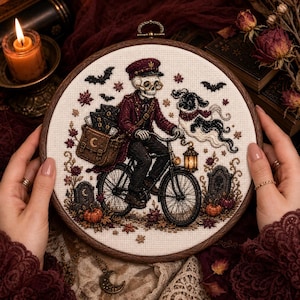 Op de afbeelding: Een ronde borduurring met een kruissteekontwerp van een skelet in een bordeauxrood uniform en pet op een fiets. Het skelet draagt een tas vol brieven, vergezeld door een spookhond, pompoenen en grafstenen.