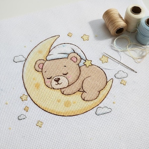 May include: Cross-stitch design featuring a sleeping teddy bear on a crescent moon, wearing a nightcap. The bear is light brown with pink accents, surrounded by yellow stars and clouds. Spools of thread and a needle are in the upper right corner.