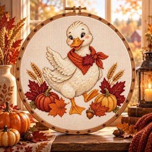 Peut inclure: Un tambour à broder circulaire en bois contenant un motif au point de croix représentant un canard blanc portant une écharpe rouge, entouré d'éléments automnaux dont des citrouilles orange, des feuilles d'érable rouges et un gland.