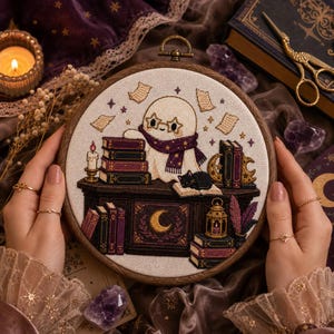 May include: A circular embroidery hoop containing a cross-stitch design of a white ghost wearing star-shaped glasses and a purple scarf. The ghost sits at a dark desk with stacks of books, a black cat, a candle, a lantern, and a crescent moon.