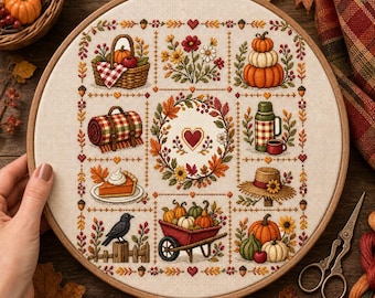 May include: A circular wooden embroidery hoop containing a cross-stitch sampler on cream fabric. The design features a central autumn wreath with a heart, surrounded by eight grid squares depicting pumpkins, a picnic basket, a thermos, and a crow.
