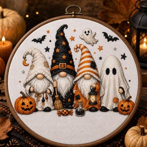 May include: A circular embroidery hoop containing a cross-stitch pattern of two gnomes, one figure in a ghost costume, and one figure in a witch hat. The design features orange pumpkins, a small lantern, a potion bottle, a candy, and a ghost.