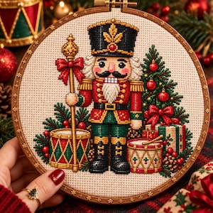 Può includere: Un ricamo a punto croce in un telaio rotondo di legno raffigurante un soldato schiaccianoci in uniforme rossa e verde, che tiene un bastone dorato. Il disegno include un albero di Natale decorato, due tamburi e regali su tessuto color crema.