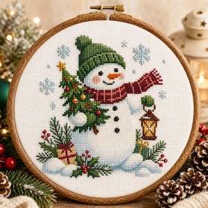 Op de afbeelding: Een kruissteekborduurwerk in een ronde houten borduurring met een sneeuwpop die een groene gebreide muts en een rood geruite sjaal draagt. De sneeuwpop houdt een versierde kerstboom en een lantaarn vast, omringd door sneeuwvlokken en hulst.