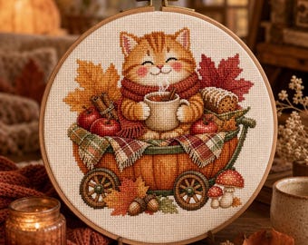 May include: A circular embroidery hoop containing a cross-stitch design of an orange tabby cat wearing a red scarf and holding a mug. The cat sits in a pumpkin-shaped cart with red apples, cinnamon sticks, autumn leaves, and mushrooms.