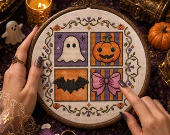 Op de afbeelding: Een ronde houten borduurring met een kruissteekpatroon verdeeld in vier vierkanten. Het ontwerp toont een wit spook op paars, een oranje pompoen, een zwarte vleermuis op oranje en een roze strik op paars-oranje gestreepte achtergrond.