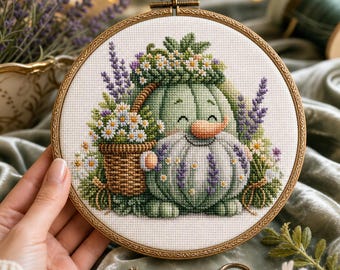 May include: A circular wooden embroidery hoop containing a cross-stitch pattern of a light green pumpkin character with a smiling face, wearing a floral crown, and holding a woven basket filled with white daisies and purple lavender sprigs.