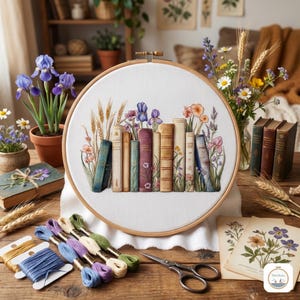 Può includere: Un telaio da ricamo a punto croce con un disegno di libri, grano e fiori. Il ricamo è su tessuto bianco, incorniciato da un telaio di legno. Sono visibili anche fili da ricamo e forbici.
