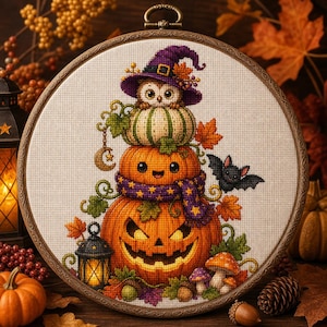Può includere: Un telaio da ricamo circolare contenente un disegno a punto croce di una pila di tre zucche. La zucca in alto presenta un gufo con un cappello da strega viola. La zucca centrale indossa una sciarpa viola e quella in basso è una zucca intagliata.