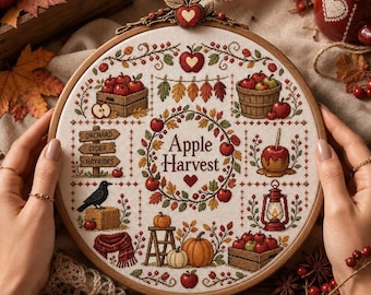May include: A round wooden embroidery hoop containing a cross-stitch design titled Apple Harvest. The pattern features apples, pumpkins, a crow, a lantern, and a signpost reading Orchard, Cider, Hayrides, all surrounded by autumn leaves.