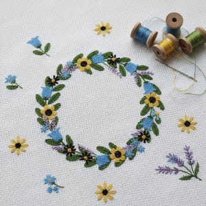 May include: A cross-stitch floral wreath with yellow sunflowers, blue bellflowers, and lavender sprigs. The wreath is surrounded by loose flowers and embroidery thread spools in blue, yellow, and green. A needle and thread are also visible.