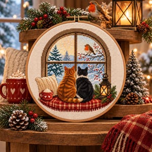 May include: A circular embroidery hoop containing a cross-stitch design of an orange tabby cat and a black cat sitting on a red plaid cushion, looking out a window at a snowy landscape with a robin on a branch and a glowing lantern nearby.