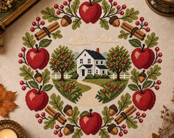 May include: A cross-stitch embroidery on light beige fabric featuring a circular wreath of red apples, green oak leaves, brown acorns, and cinnamon sticks surrounding a central scene of a white house with a black roof and two apple trees.