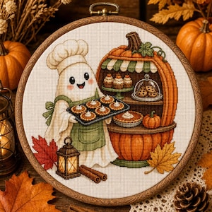 May include: A cross-stitch embroidery in a round wooden hoop featuring a white ghost wearing a chef hat and green apron, holding a tray of small pies. Next to the ghost is a large pumpkin-shaped bakery stand with cupcakes, cookies, and a pie.