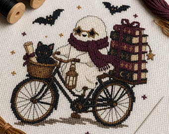 Op de afbeelding: Een kruissteekpatroon op witte stof met een witte geest met stervormige bril en een donkerpaarse sjaal op een fiets. De fiets heeft een mandje met een zwarte kat en een stapel boeken achterop, versierd met vleermuizen en sterren.